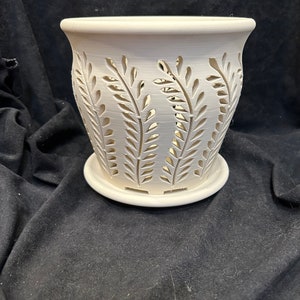May include: A white ceramic planter with a matching saucer. The planter features a cut-out leaf design around the sides. The pot has a slightly flared top and is suitable for indoor or outdoor use.