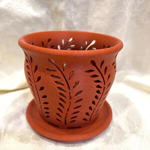 May include: A terracotta-colored ceramic planter with a speckled finish and a matching saucer. The pot features a repeating cut-out leaf pattern around its exterior, allowing for ventilation. The design includes a wide, flared rim and a circular base.