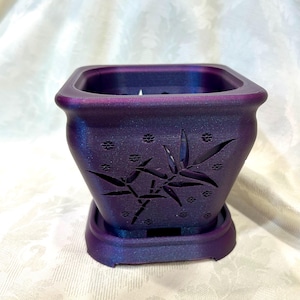 May include: A square, purple planter with a matching saucer. The planter has a floral design cut into the sides and a textured finish. The planter is designed to hold a small plant. The planter is approximately 5 inches wide and 4 inches tall.