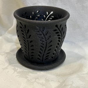 May include: Black ceramic planter with a matching saucer. The planter features a cut-out leaf design around the sides. The interior is dark, and the rim has a slightly textured appearance. The planter is designed for indoor or outdoor use.
