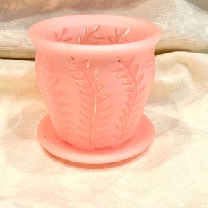 May include: A light pink, frosted glass planter with a matching saucer. The planter has a cylindrical shape with a flared rim and is decorated with a repeating leaf pattern. The saucer is round and fits snugly beneath the planter.