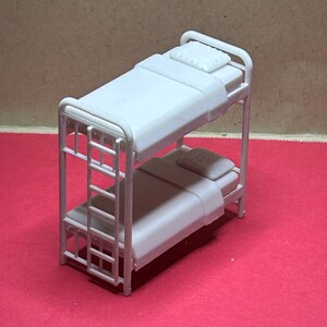 May include: A miniature white bunk bed with a ladder, two mattresses, and two pillows. The bed frame is made of thin metal bars. The top bunk is slightly higher than the bottom bunk. The bed is on a red surface.