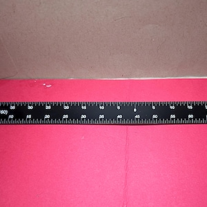 May include: A black ruler with white markings and numbers, measuring up to 75 units. The ruler is placed on a red surface, with a section of a brown surface visible in the background. The ruler has "N Scale (1:160)" printed on it.