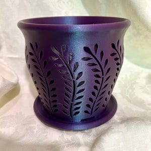 May include: A purple planter with a leaf design cut into the sides. The planter has a round base and a flared top. The color shifts from purple to blue in the light. The planter is designed for indoor or outdoor use.
