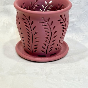 May include: A pink ceramic planter with a decorative leaf pattern. The planter has drainage holes and a matching saucer.