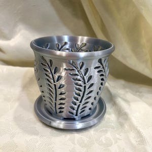 May include: A silver-colored, decorative planter with a matching saucer. The planter features a cylindrical shape with cut-out leaf designs. The planter is designed to hold small plants or flowers, adding a touch of elegance to any space.