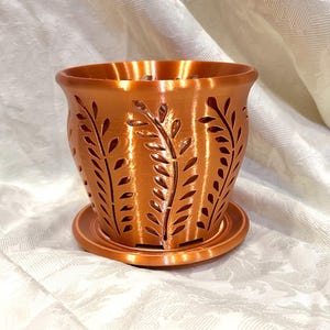 May include: A copper-colored planter with a matching saucer. The planter features a cylindrical shape with cut-out leaf designs. The planter is approximately 5 inches in diameter and 4 inches tall, suitable for small plants or succulents.