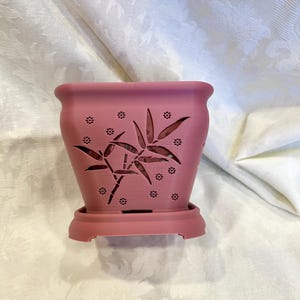 May include: A pink plastic planter with a cutout design of bamboo leaves and small flowers. The planter has a drainage tray.