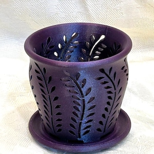 May include: A deep purple, shimmering plant pot with a matching circular saucer. The pot features a repeating cut-out pattern of vertical fern-like leaves that wrap around the exterior, allowing light to pass through the openings in the wall.