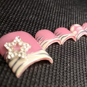 Puede incluir: Uñas postizas rosas con un adorno de pedrería en forma de estrella y diseño de rayas de cebra. Las uñas tienen un acabado brillante y están dispuestas sobre una superficie negra texturizada. Las puntas de las uñas tienen un diseño de rayas de cebra blancas y negras.