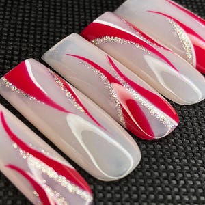 Op de afbeelding: Een set van zes lange, ovaalvormige kunstnagels. Elke nagel heeft een heldere basis met een rood, gebogen ontwerp en een zilveren glitteraccent. De nagels zijn gerangschikt op een zwart oppervlak.