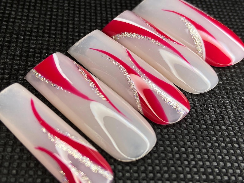 Op de afbeelding: Een set van zes lange, ovaalvormige kunstnagels. Elke nagel heeft een heldere basis met een rood, gebogen ontwerp en een zilveren glitteraccent. De nagels zijn gerangschikt op een zwart oppervlak.