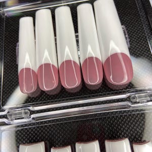 May include: A set of ten white false nails with a pink tip. The nails are arranged in two rows in a clear plastic container.