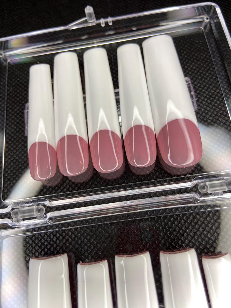 May include: A set of ten white false nails with a pink tip. The nails are arranged in two rows in a clear plastic container.