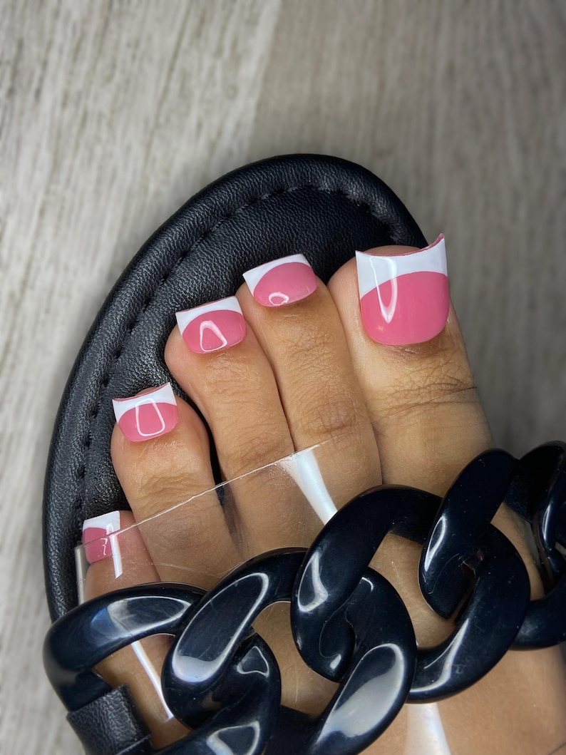 May include: Close-up of a foot with pink and white French tip toenails. The nails are painted with a glossy finish. The foot is in a black sandal with a clear strap and a chunky black chain detail.