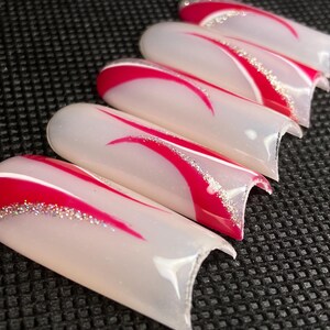 Op de afbeelding: Een set van vijf kunstnagels. Elke nagel is wit met een rood gebogen ontwerp en een zilveren glitteraccent. De nagels zijn diagonaal geplaatst tegen een donkere, getextureerde achtergrond.