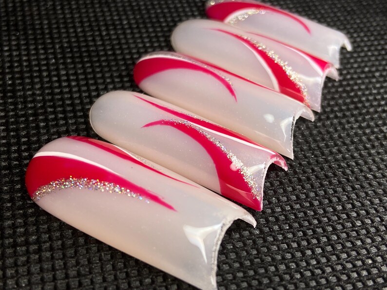 Op de afbeelding: Een set van vijf kunstnagels. Elke nagel is wit met een rood gebogen ontwerp en een zilveren glitteraccent. De nagels zijn diagonaal geplaatst tegen een donkere, getextureerde achtergrond.
