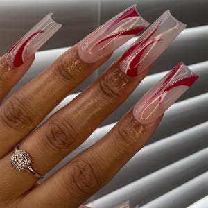 Op de afbeelding: Lange, vierkante acrylnagels met een heldere basis, rode uiteinden en een rood en zilver wervelontwerp. Een zilveren ring met een vierkante, diamantachtige steen zit op de ringvinger. De nagels zijn een modieus accessoire.