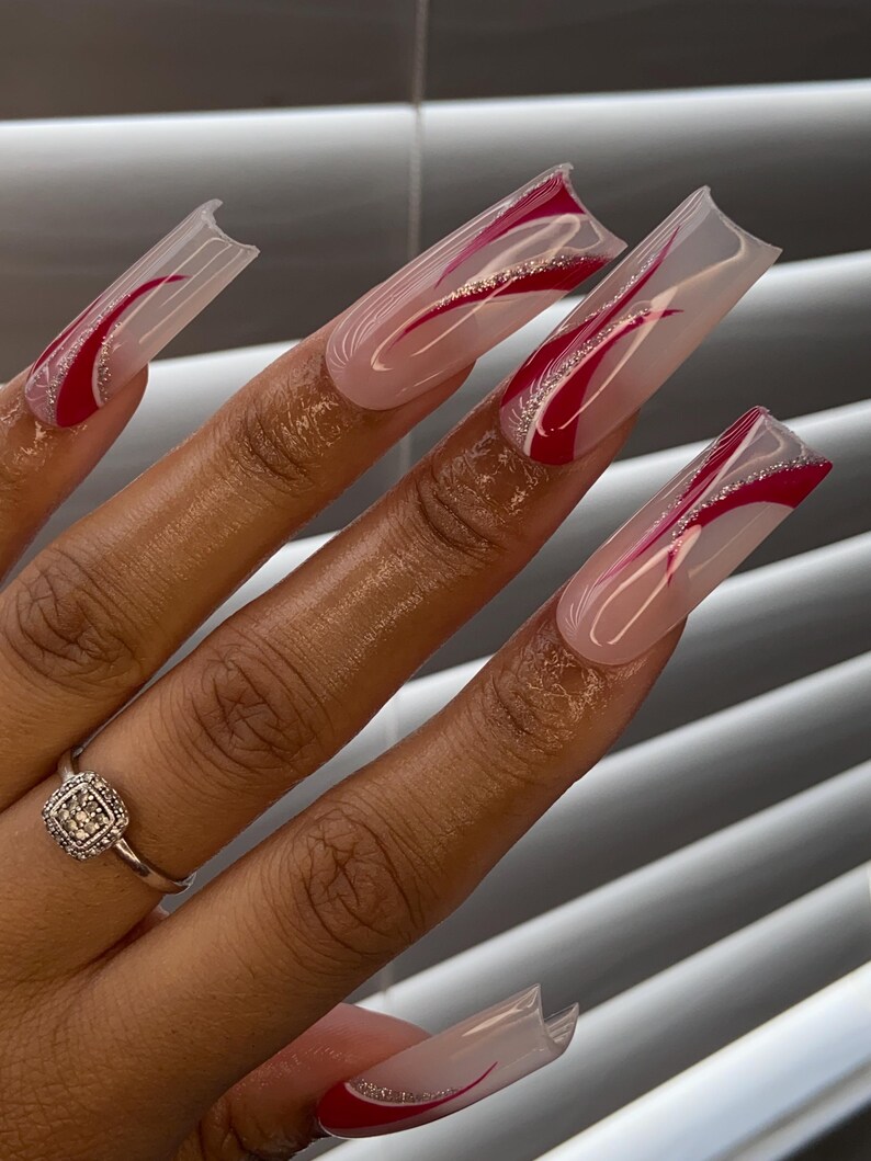 Op de afbeelding: Lange, vierkante acrylnagels met een heldere basis, rode uiteinden en een rood en zilver wervelontwerp. Een zilveren ring met een vierkante, diamantachtige steen zit op de ringvinger. De nagels zijn een modieus accessoire.
