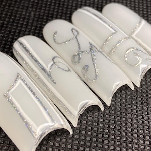 May include: Five white artificial nails with silver glitter accents. The nails feature various designs, including a rectangular frame, stripes, and cursive letters. The nails are arranged on a textured black surface.