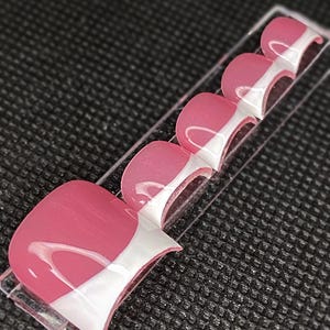 May include: A set of pink and white press-on toenails. Each nail has a glossy finish with a pink base and a white curved design. The nails are arranged on a clear plastic strip against a textured black background.