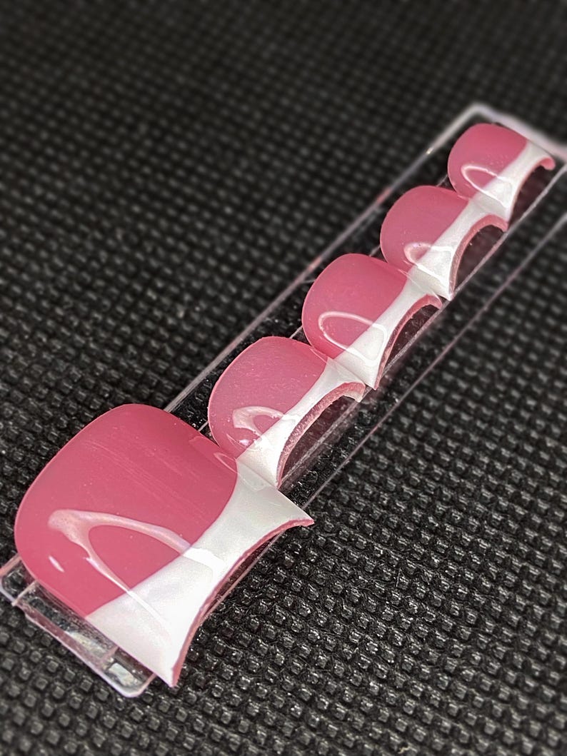 May include: A set of pink and white press-on toenails. Each nail has a glossy finish with a pink base and a white curved design. The nails are arranged on a clear plastic strip against a textured black background.