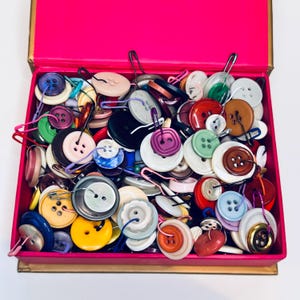 May include: A collection of colorful buttons in a pink box. The buttons vary in size, shape, and color, including red, blue, yellow, and green. Many buttons have safety pins attached. The box has a gold-colored trim.