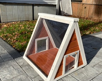 May include: A-frame style pet house with a clear plastic roof and a wooden door. The house is painted white with a red wooden interior. The house is sitting on a gray concrete patio.