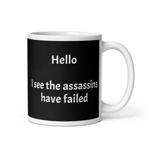 May include: White ceramic mug with a black design. The design says "Hello I see the assassins have failed".
