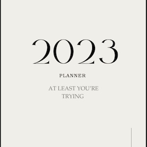 May include: A white planner with the year 2023 printed in black. The text "Planner" is printed below the year, and the text "At least you're trying" is printed below that.
