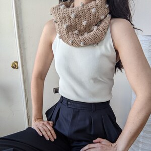 May include: A brown crocheted cowl with beige pom-poms. The cowl is worn by a person wearing a white tank top and black pants.