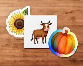 Puede incluir: Un conjunto de pegatinas con un girasol, una vaca y una calabaza. El girasol tiene pétalos amarillos y un centro marrón. La vaca es marrón y blanca. La calabaza es naranja y verde.