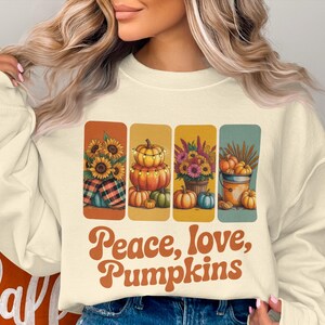 May include: A cream-coloured sweatshirt with a fall-themed graphic. The graphic features four rectangular panels with images of sunflowers, pumpkins, and the text "Peace, love, Pumpkins" in a retro font. The design evokes autumn.