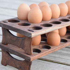 May include: A two-tiered, wooden egg holder filled with brown eggs. The dark brown holder has circular cutouts to hold the eggs. The eggs are a light brown color and are evenly spaced in the holder. The wooden holder is on a light-colored wooden surface.