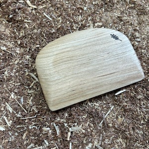 May include: A light-colored, wooden dough scraper with a curved edge and a small, dark leaf design. The scraper is resting on a bed of wood shavings, showcasing its natural wood grain and craftsmanship. This kitchen tool is designed for baking and food preparation.