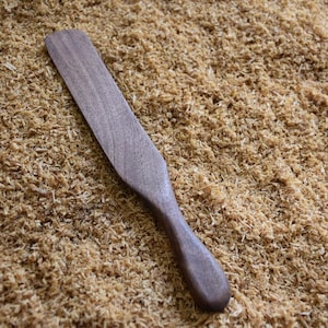 May include: A wooden spatula with a curved handle, lying on a bed of wood shavings. The spatula is a dark brown color and appears to be handmade. The wood shavings are light brown and create a textured background.