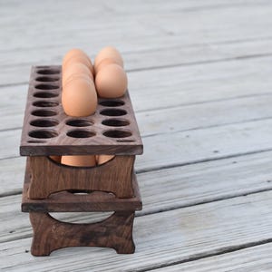 Pode incluir: Um suporte de madeira para ovos de dois n&iacute;veis, cada um com recortes circulares para ovos. V&aacute;rios ovos castanhos s&atilde;o colocados no n&iacute;vel superior. O suporte &eacute; feito de madeira escura e assenta numa superf&iacute;cie de madeira clara.
