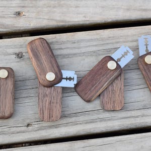 May include: A collection of wooden razor blade holders with brass accents. The holders are rectangular with rounded edges and a hinged design to hold the blades. The wood has a rich, dark brown color, and the blades are silver.