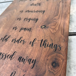 May include: A rectangular dark brown wooden board with a natural wood grain and visible knots. The board features deeply engraved black text in a cursive script that reads: He will wipe every tear from their eyes. There will be no more death or mourning or crying or pain, for the old order of things has passed away. Revelation 21:4.