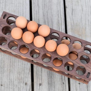 Pode incluir: Um suporte de madeira para ovos de dois n&iacute;veis, cada um com recortes circulares para os ovos. O suporte est&aacute; cheio de ovos castanhos. A madeira tem um acabamento castanho escuro, e o suporte est&aacute; sobre uma superf&iacute;cie de madeira cinza claro.