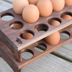 Pode incluir: Um porta-ovos de madeira de dois n&iacute;veis, tingido de castanho escuro, cheio de ovos castanhos. O suporte tem recortes circulares para segurar os ovos e est&aacute; sobre uma superf&iacute;cie de madeira. Um acess&oacute;rio de cozinha.