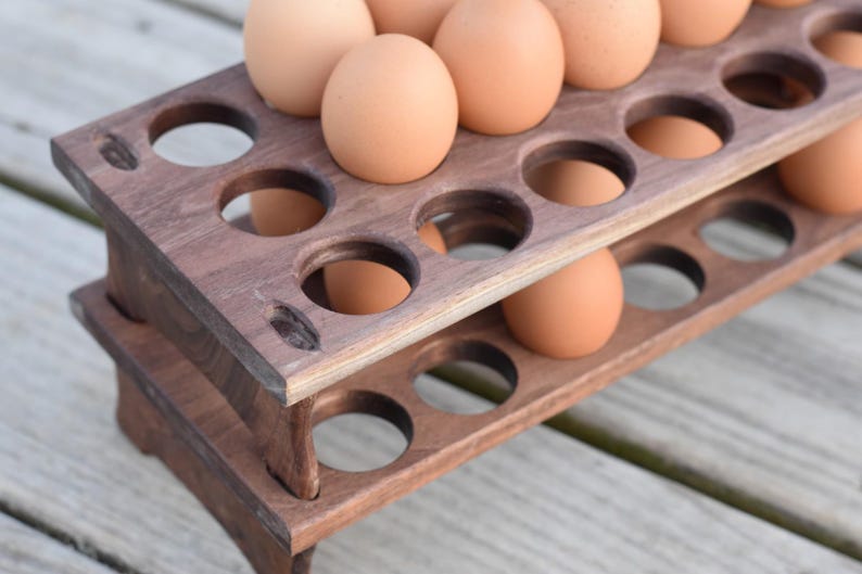 Pode incluir: Um porta-ovos de madeira de dois n&iacute;veis, tingido de castanho escuro, cheio de ovos castanhos. O suporte tem recortes circulares para segurar os ovos e est&aacute; sobre uma superf&iacute;cie de madeira. Um acess&oacute;rio de cozinha.