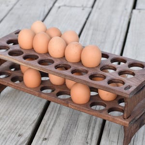 Pode incluir: Um suporte para ovos de madeira castanha escura de dois n&iacute;veis, cheio de ovos castanhos. O suporte tem recortes circulares para segurar os ovos e est&aacute; sobre uma superf&iacute;cie de madeira. &Eacute; um artigo de armazenamento de cozinha.