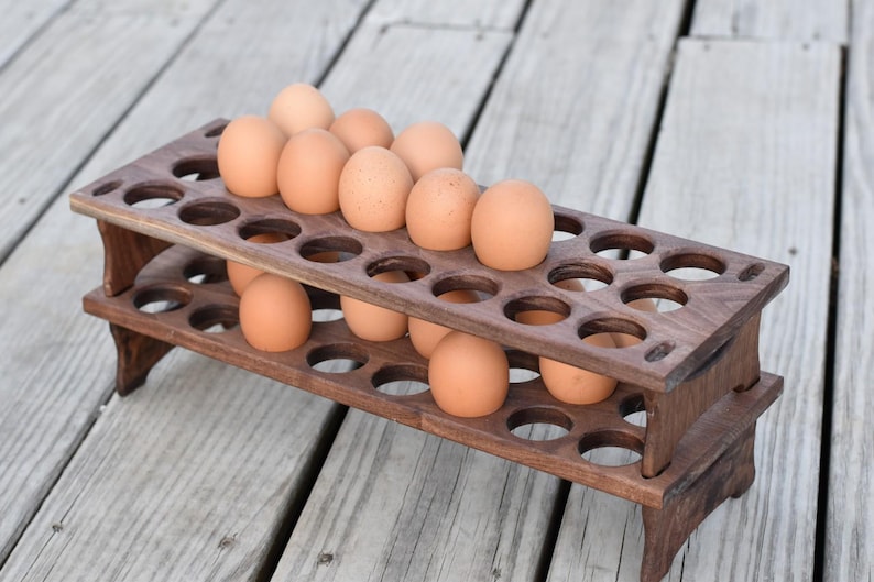 Pode incluir: Um suporte para ovos de madeira castanha escura de dois n&iacute;veis, cheio de ovos castanhos. O suporte tem recortes circulares para segurar os ovos e est&aacute; sobre uma superf&iacute;cie de madeira. &Eacute; um artigo de armazenamento de cozinha.