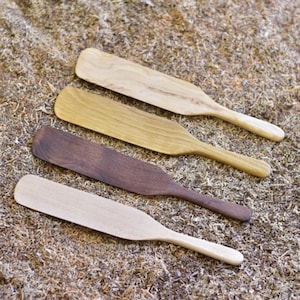May include: Four wooden spatulas arranged diagonally, each featuring a distinct wood grain and color ranging from light beige to dark brown. These kitchen utensils have a flat, wide blade and a rounded handle, showcasing natural wood textures.