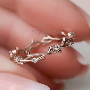 Könnte beinhalten: Ein zarter Goldring mit einem Zweigdesign und kleinen, klaren Steinen. Der Ring befindet sich an einem Finger mit rosafarbenem Nagellack.