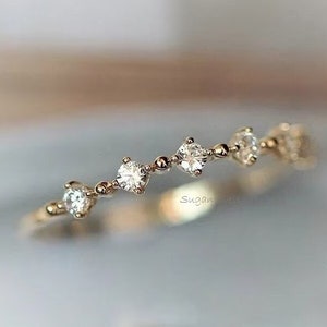 Pode incluir: Um delicado anel de banda de ouro com uma fila de pequenas pedras preciosas transparentes. O anel tem um design simples e elegante.