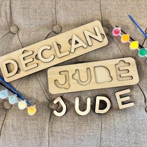 May include: Two light-colored wooden name puzzles featuring the names DECLAN and JUDE, with the individual wooden letters for JUDE placed below. Two sets of small plastic paint pots in various colors and two paintbrushes are included for crafting.