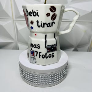 Puede incluir: Taza de café de cerámica blanca con asa, decorada con granos de café, estrellas y las palabras "Debi tirar mas fotos". La taza tiene una cuchara dorada y se apoya en una base blanca con pedrería plateada.