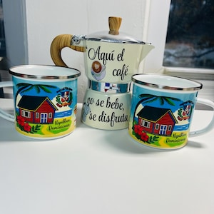 Puede incluir: Un juego de café con una cafetera italiana crema y dos tazas esmaltadas a juego. Las tazas y la cafetera tienen el texto "Aquí el café, no se bebe, se disfruta". Las tazas tienen un diseño azul y amarillo con una casa y el texto "República Dominicana".
